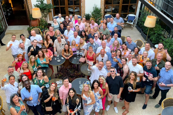 A group of 85 people stood in an outdoor bar setting, looking upwards at the camera and posing happily for a photograph.