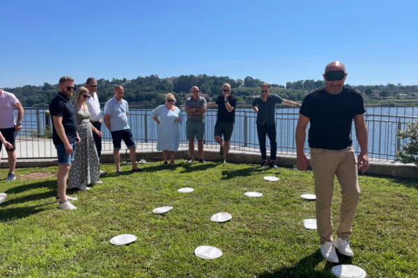 Group watching as blindfolded man coordinates through numbered paper plates on grass, with river, trees, and clear blue sky in the background.